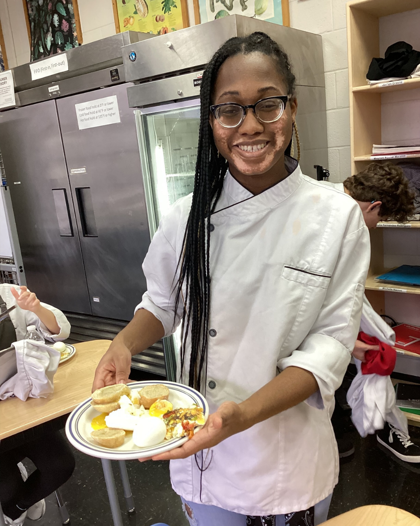 A student dressed in a chef jacket smile for her photo holding the plate with eggs she made inside the kitchen. 