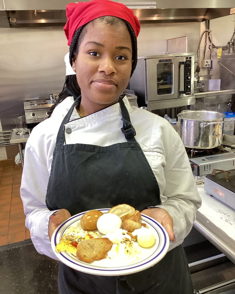 A student wears a chef hat and apron smiles with her creation of eggs on a plate inside the kitchen. 