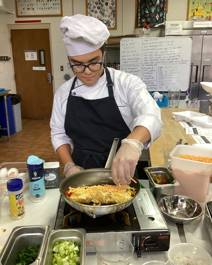 A student wearing a chef hat and apron cooks his omelet on a stove inside the kitchen.