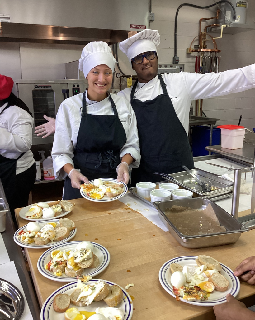 Two students dressed in a chef hat and apron smile for a photo inside the kitchen, with six plates of egg creations on the table inside the kitchen.