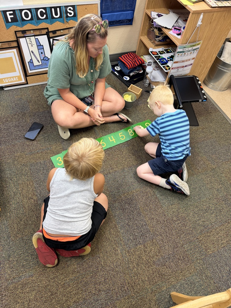 A ELC teacher sits with two ELC students learning how to count with green number cards on the floor inside the classroom.