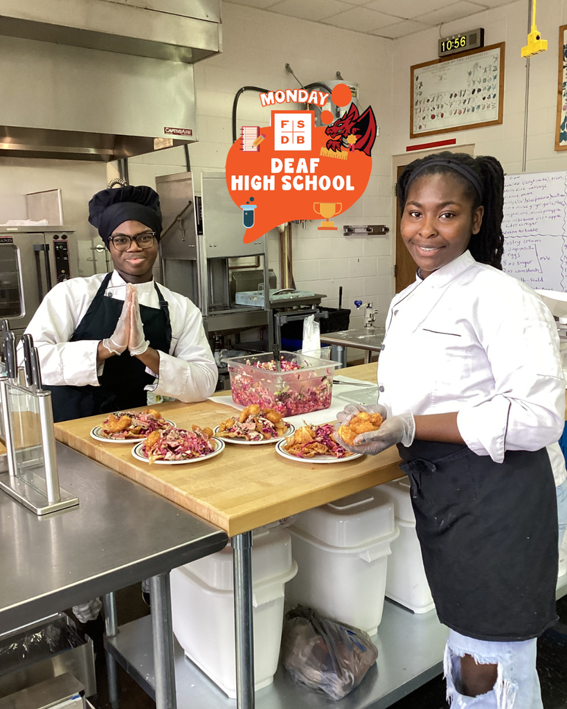 Two culinary arts students wear black and white chef gear and smile with the plates inside the kitchen.