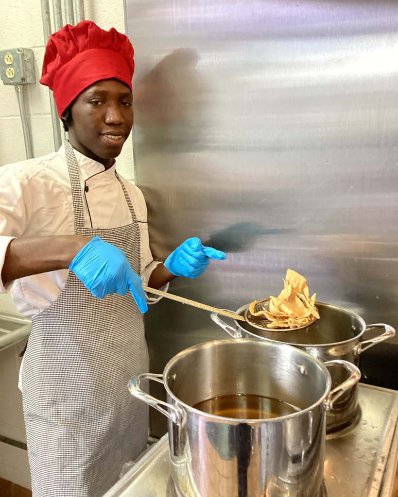 A student smiles and points to the food he is making on the stove inside the kitchen.
