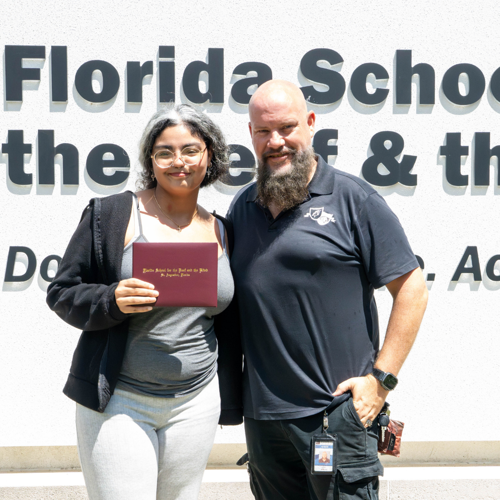 Jaylene holds her diploma and smiles with a staff member by the FSDB sign.