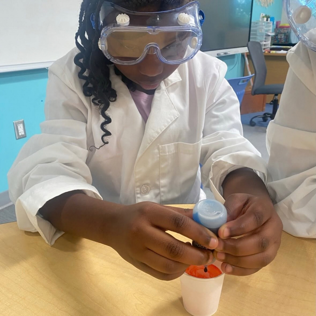 A close up photo of a student dropping blue drops into the red water inside the cup.