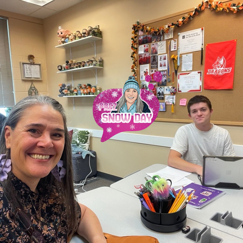 President Snow sits at a classroom table in the foreground while Logan sits across the table with a laptop, notebook, and papers. Behind Logan, shelves filled with collectible figures and a bulletin board covered with photos, certificates, and a red Dragons banner decorate the classroom wall.