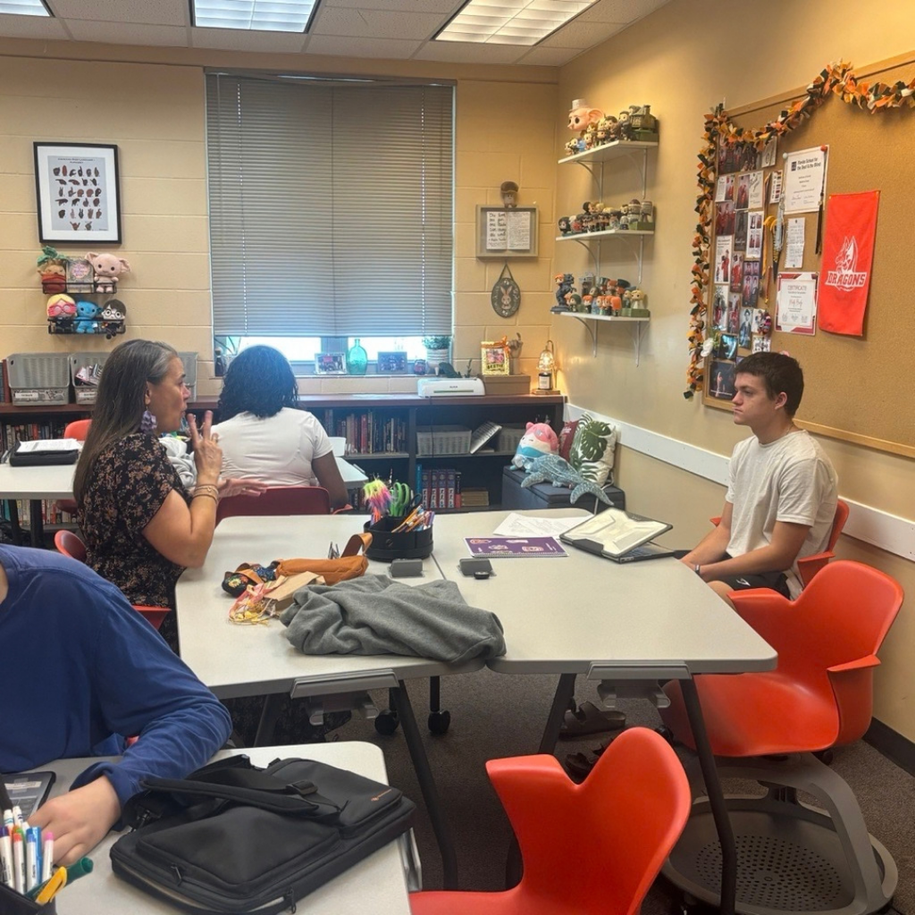 President Snow and Logan sit across from one another at a large classroom table showing President Snow answering  questions. The classroom contains bookshelves, student work, display shelves with collectibles, a large window with closed blinds, and colorful orange chairs arranged around the room.