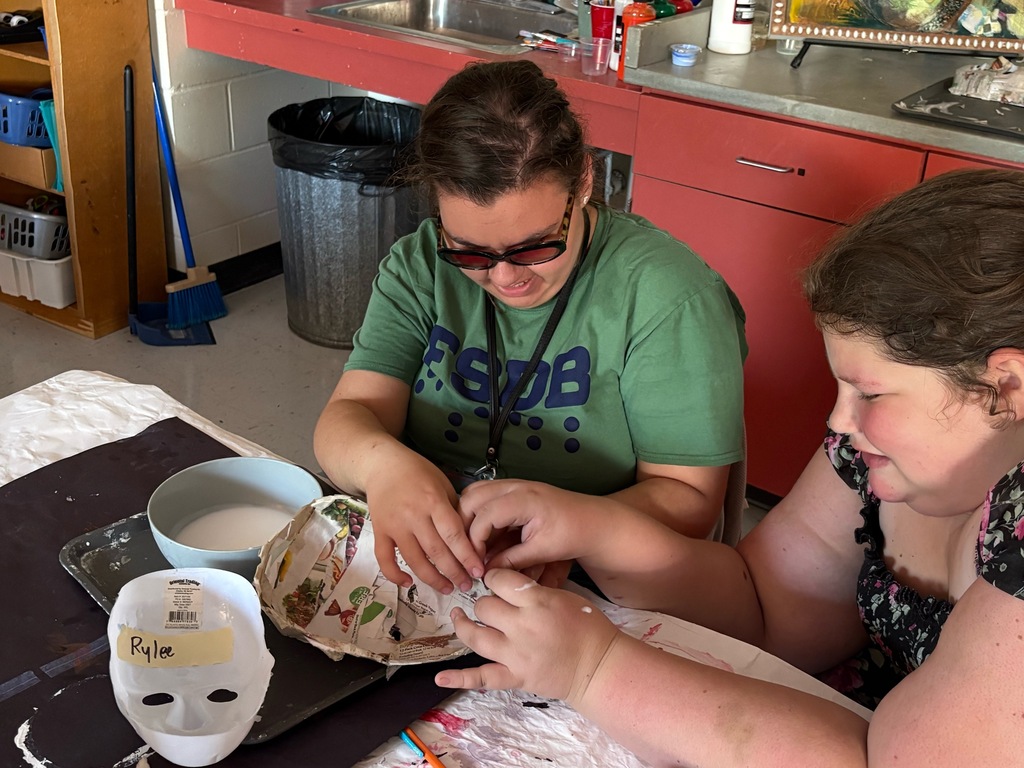 Rylee and Breanna smile while applying strips of paper to a paper mache mask.