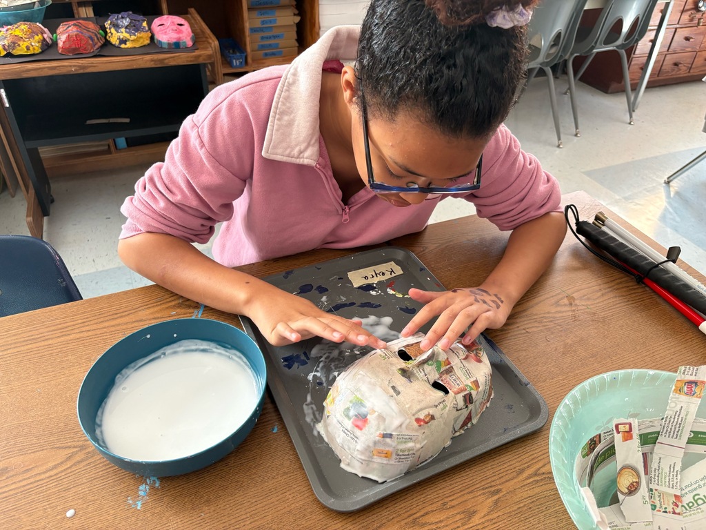 Keyra is applying a layer of glue paste over a paper mache mask, smoothing the surface with her hand.