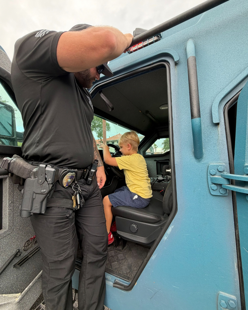 A ELC student is sitting inside the truck with a police officer watching over him.