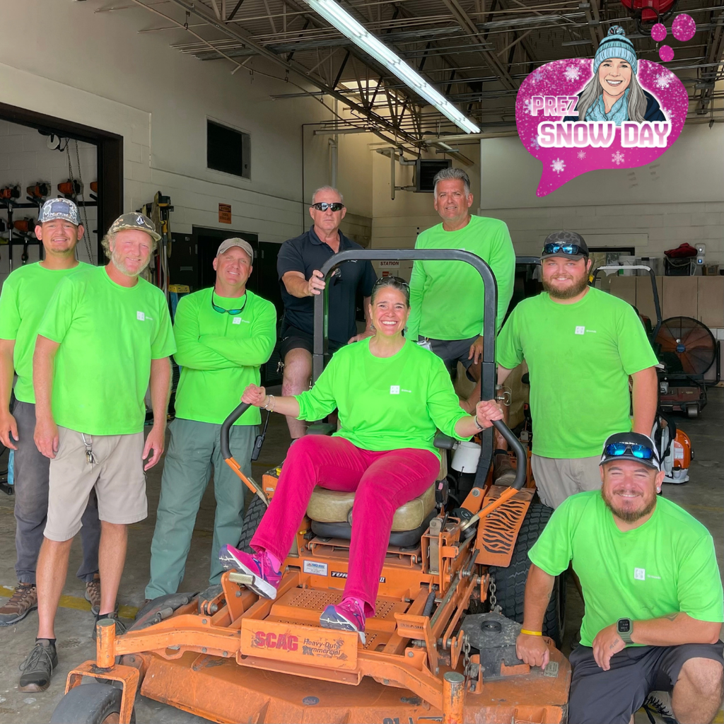Photo Description: President Snow, grinning broadly, sits on an orange commercial lawn mower positioned in the center of a maintenance garage. She is wearing bright pink pants and matching pink athletic shoes. Members of the grounds crew, dressed in matching bright green work shirts, stand or kneel around the mower, while their supervisor, wearing dark navy, joins the group as well. Lawn care equipment, tools, and the open garage space are visible in the background.