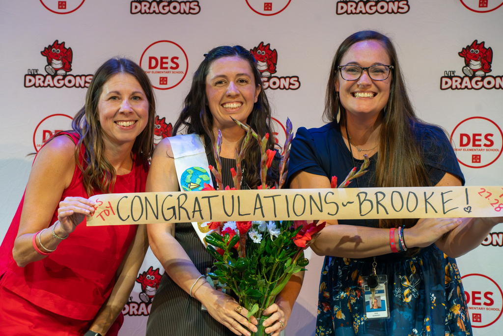 Brooke smiles with the 2026-2027 winner and Assistant Principal of DES with flowers and a sign that says "Congratulations Brooke!"