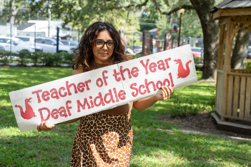 Alicia smiles outside holding the sign that says "Teacher of the Year, Deaf Middle School"