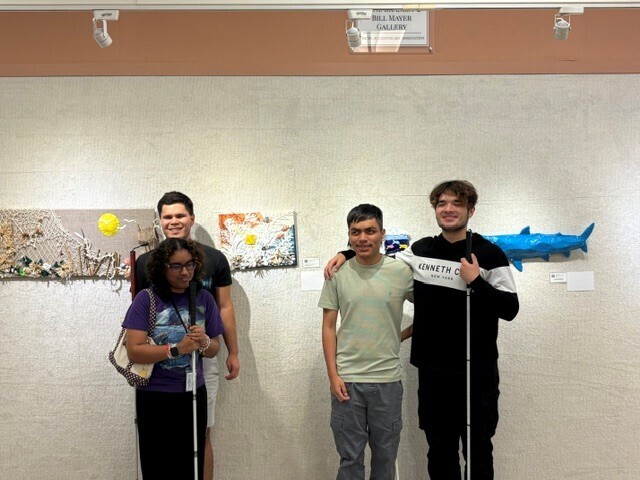 Alina, Sam, Kevin, and Aref smile as they stand in from of ocean themed canvases and shark sculpture.