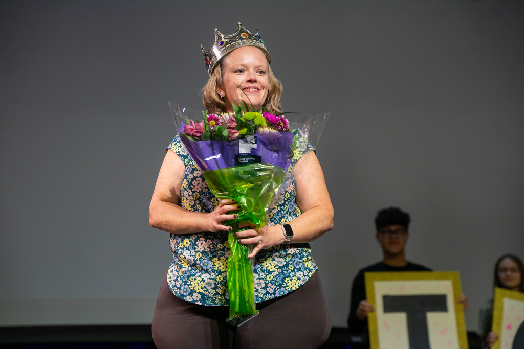 Brianna smiles with a crown on her head and holds flowers on stage in Kirk.