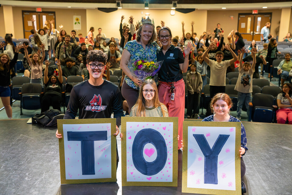 Three students smile with the TOY sign with Brianna and Jay stand behind them and students cheering in the background.