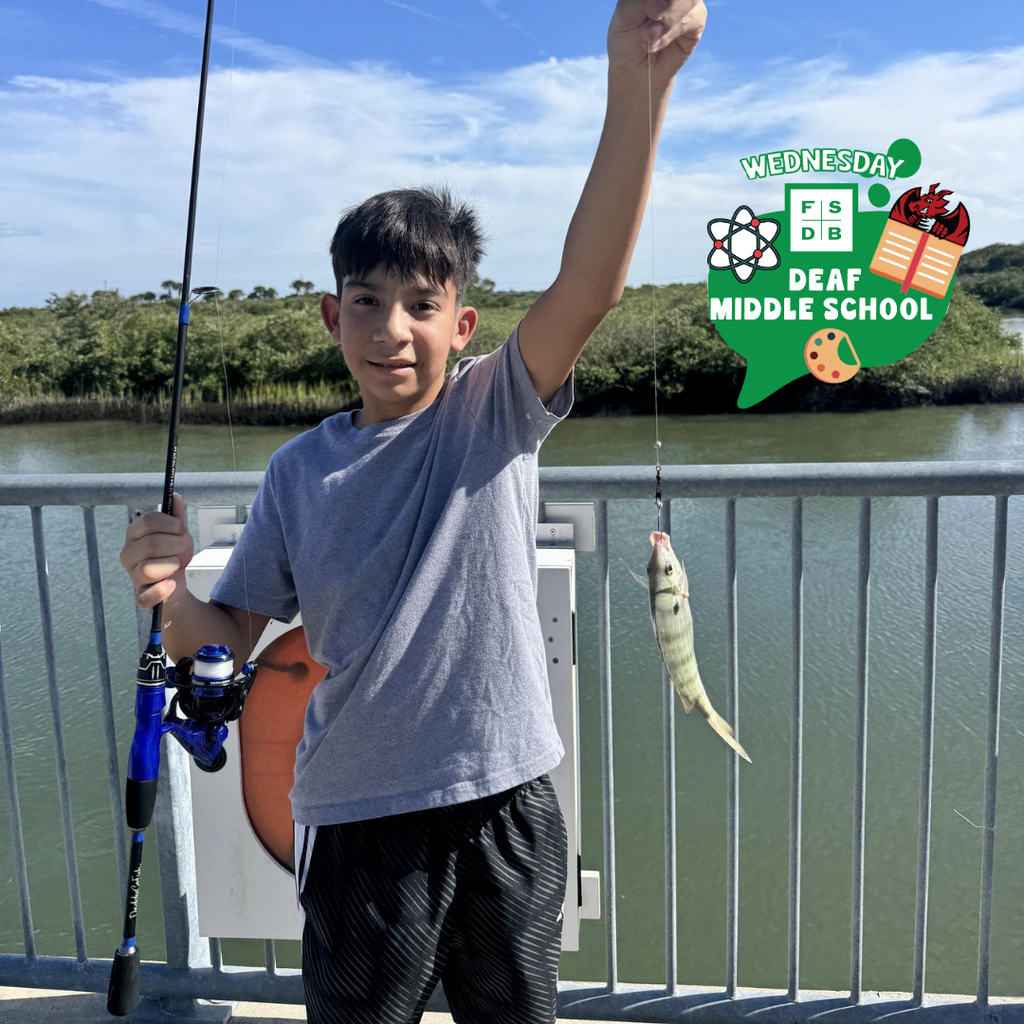Student holds a rod and fish on the end outside by the water. 