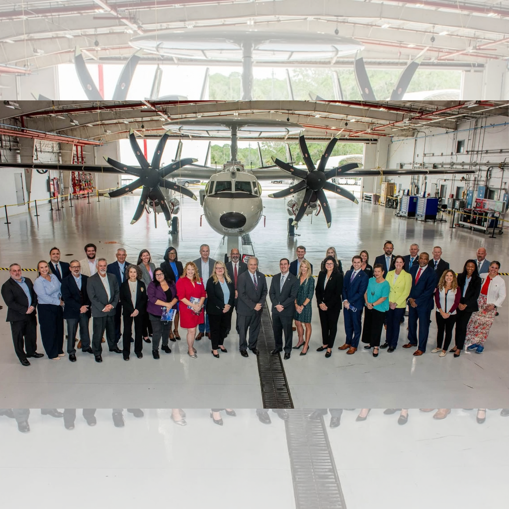 Members of Northrop Grumman, the Florida Chamber of Commerce, the St. Johns County Chamber of Commerce, and other visitors pose for a group photo inside an aircraft hangar. The group stands in front of an E-2D Advanced Hawkeye aircraft, with its distinctive radar dome and large propellers prominently displayed behind them. President Snow is standing in the front row on the far right side of the group.