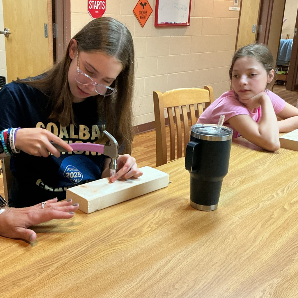 Close up of a student hammering a screw into wood and a student watching her.