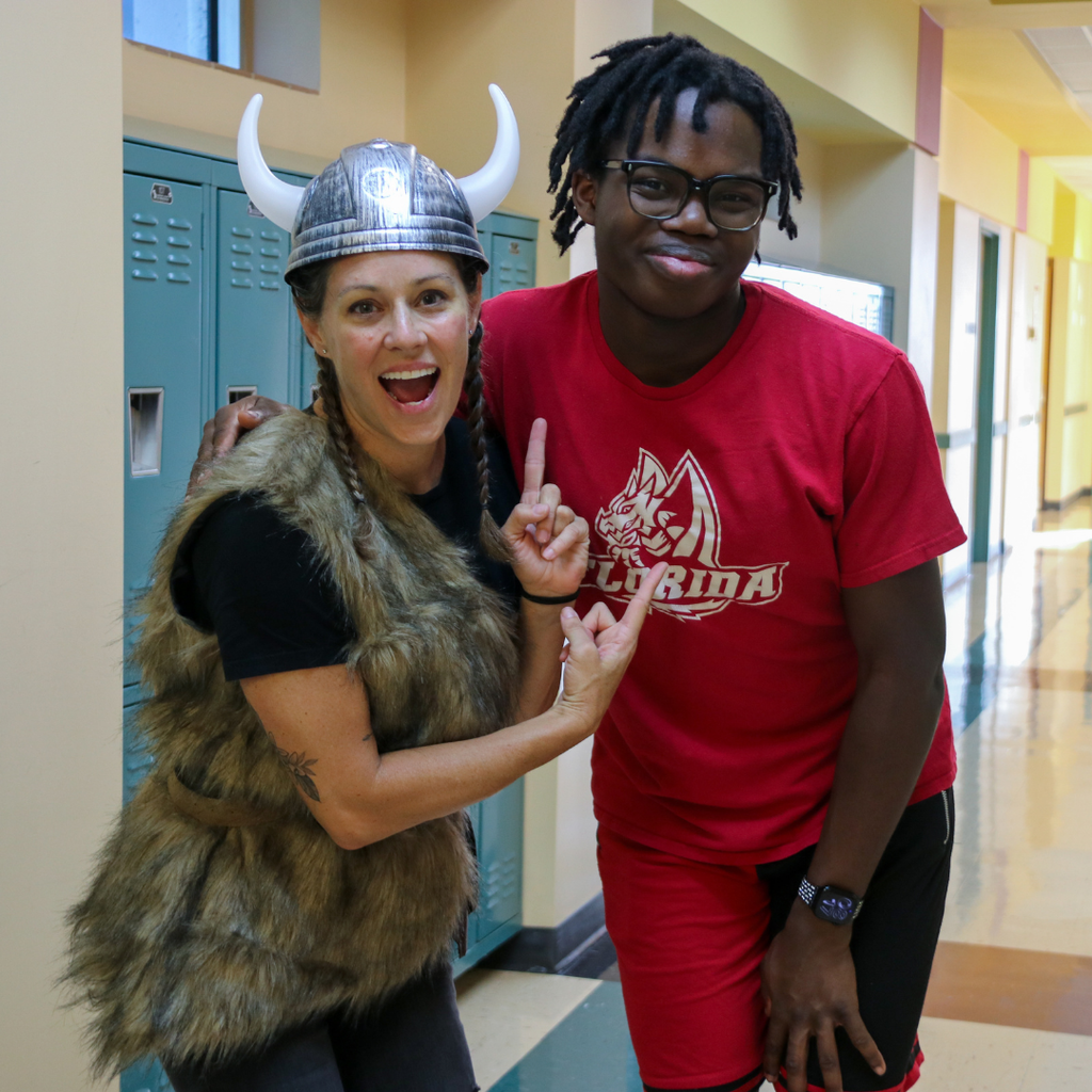 A staff member dressed like a viking and student wearing red FSDB dragons clothes smile for a photo in the hallway of Pope.
