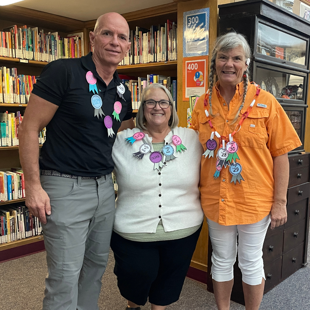 Three staff dressed in black, white and orange with a homemade necklace around them of each class name written on the ribbon inside the library.