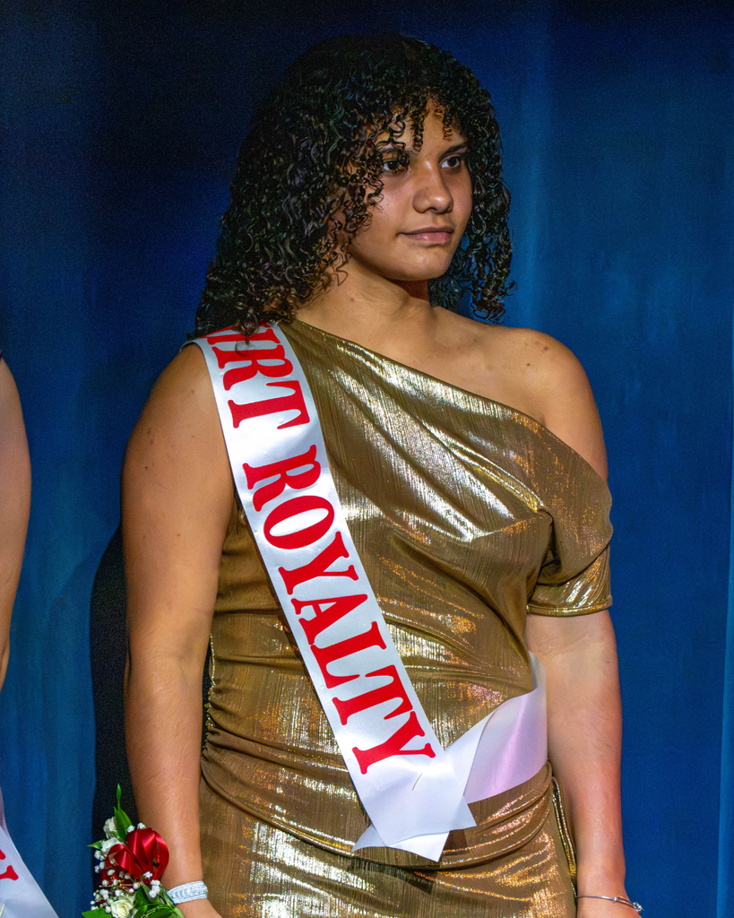 A senior girl smiles on stage in a gold dress and sash that says "Court Royalty" 