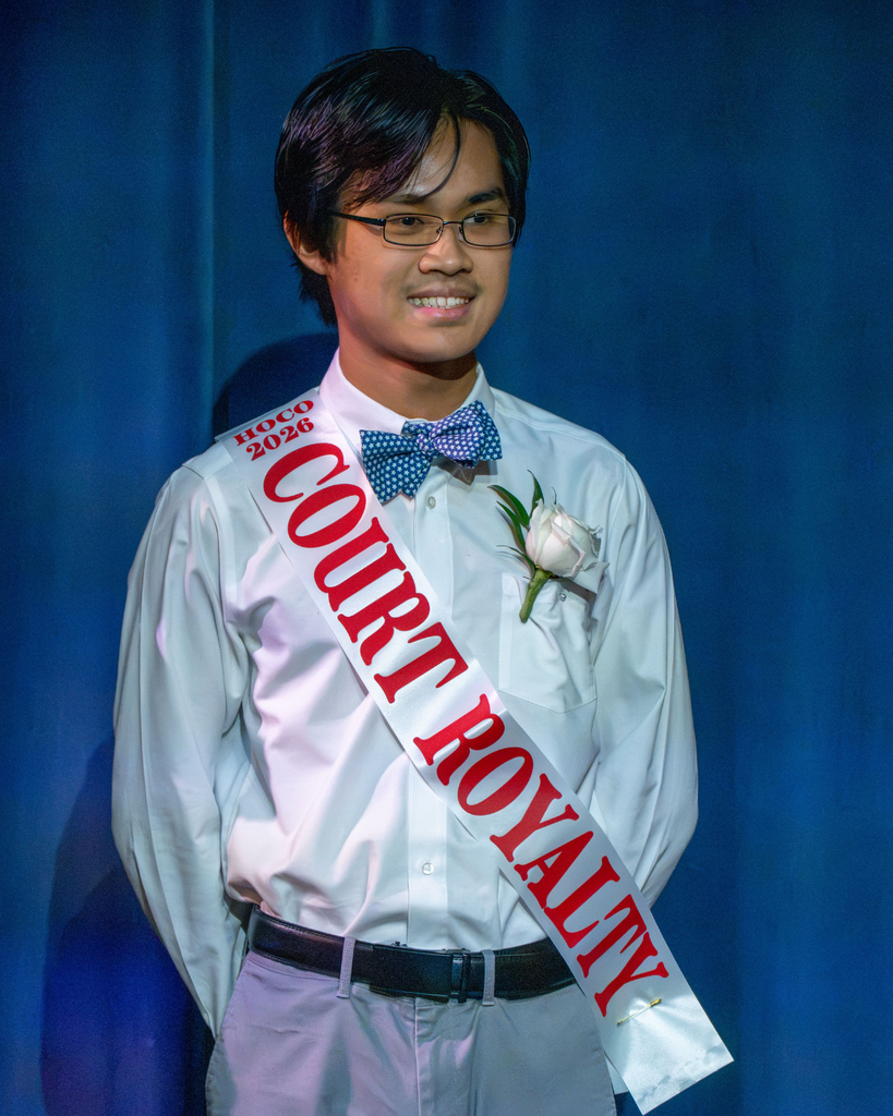 A senior smiles in a white collar shirt and a sash that says "Court royalty" on stage. 