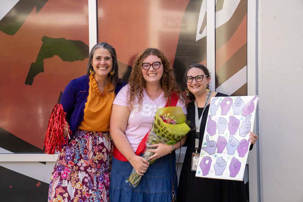 President Snow, Lexi Griffin, and DHS Assistant Principal Terri Samson stand together in front of a large graphic of the FSDB mascot outside the gym. Lexi holds a bouquet of colorful flowers and wears a red ROY sash. President Snow on the left holds red metallic pom-poms. Ms. Samson on the right holds a large poster-size card decorated with illustrations of pink and purple hands forming handshapes that spell out "Congratulations," surrounded by colorful stars. All three face the camera smiling brightly.