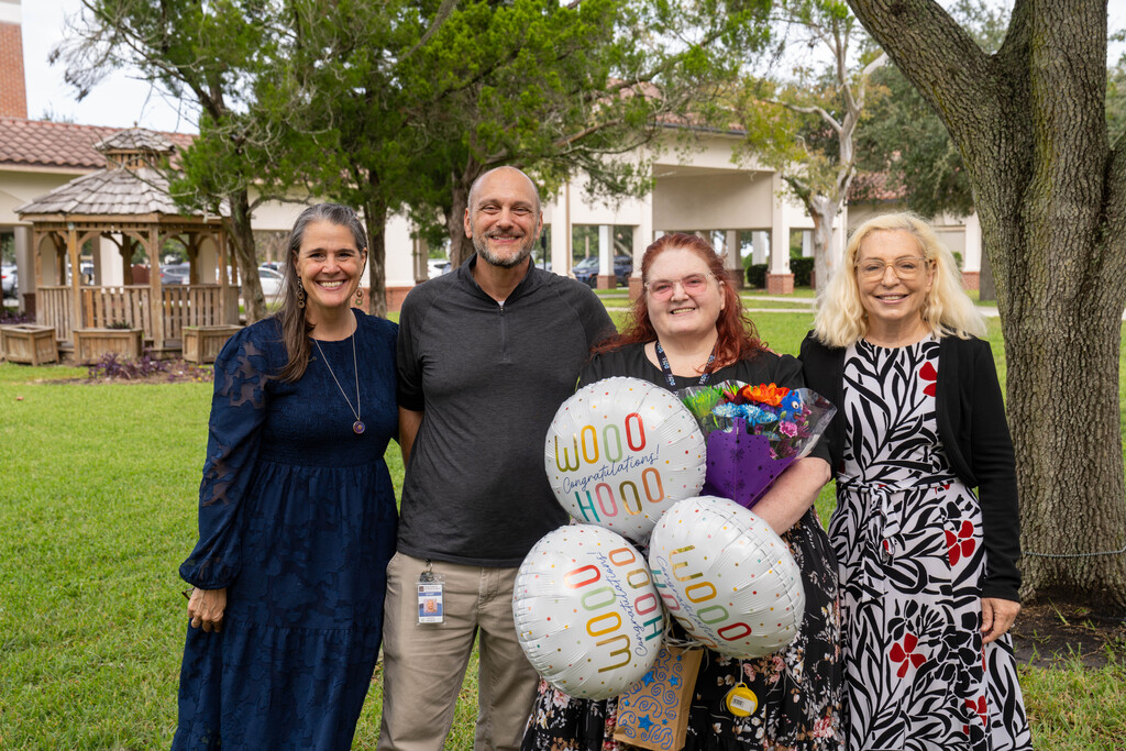 President Snow, BHS Assistant Principal Justin Cosgrove, Leslie Costello, and Principal Carol Bogue stand together, smiling outdoors on the FSDB campus beneath a large tree. Ms. Costello holds a colorful bouquet of flowers and several white balloons printed with “Woo hoo!” and “Congratulations!” A grassy courtyard, trees, a wooden gazebo, and campus buildings with red-tile roofs are visible in the background.