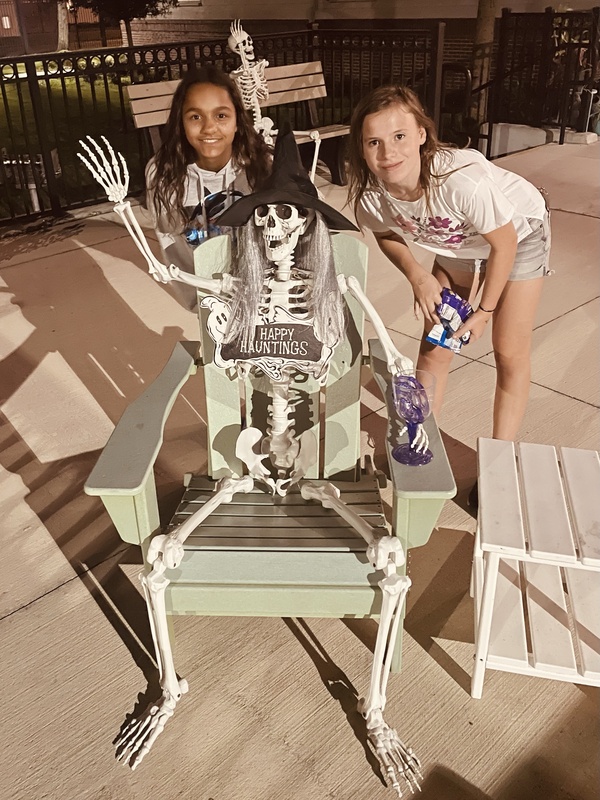 Two girls posing with a Halloween skeleton sitting in an outdoor chair.