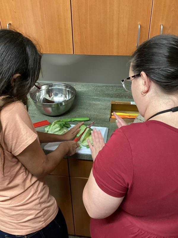 Girl cutting lettuce in the kitchen as a RI looks on.
