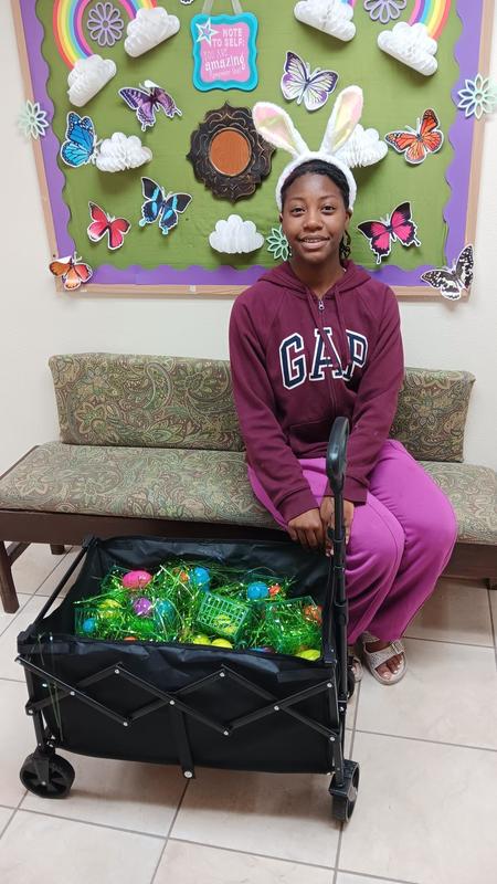 Student sitting down on bench, smiling with carriage full of Easter eggs and bunny ears on