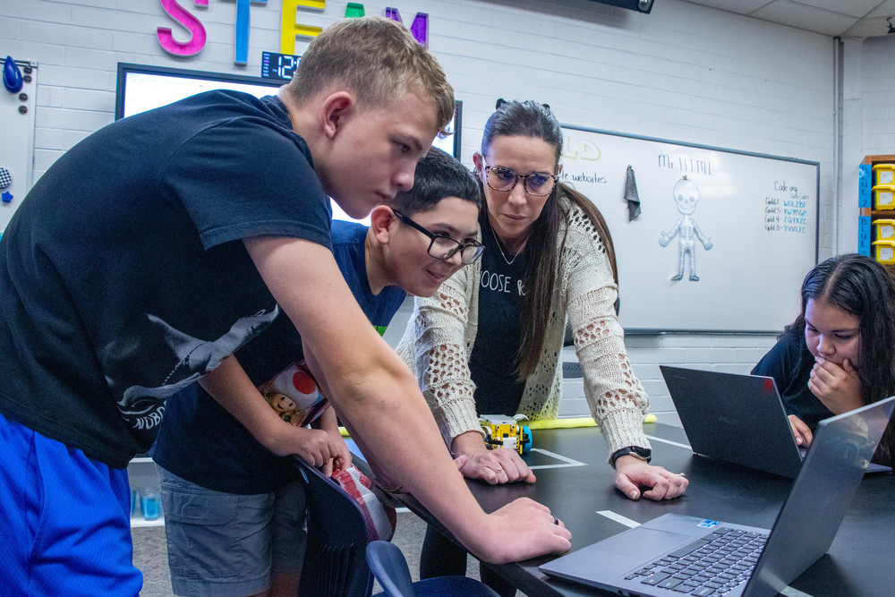 Teacher and three students lean over a laptop in a STEAM classroom, collaborating on a project. Laptops and a small robot sit on the table with a whiteboard in the background.