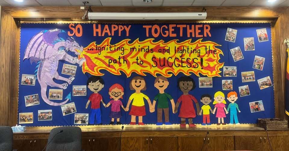 Bulletin board display in the district board room created by Homer Davis. The board reads ‘So Happy Together… igniting minds and lighting the path to success!’ with a purple dragon breathing fire across the center.