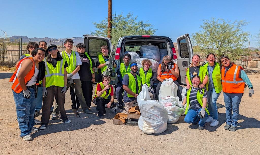 Group of Sentinel Peak High School students and staff in safety vests pose by an open van after a roadside cleanup. Large bags of collected trash and grabbers sit in front, with desert trees and mountains in the  background.