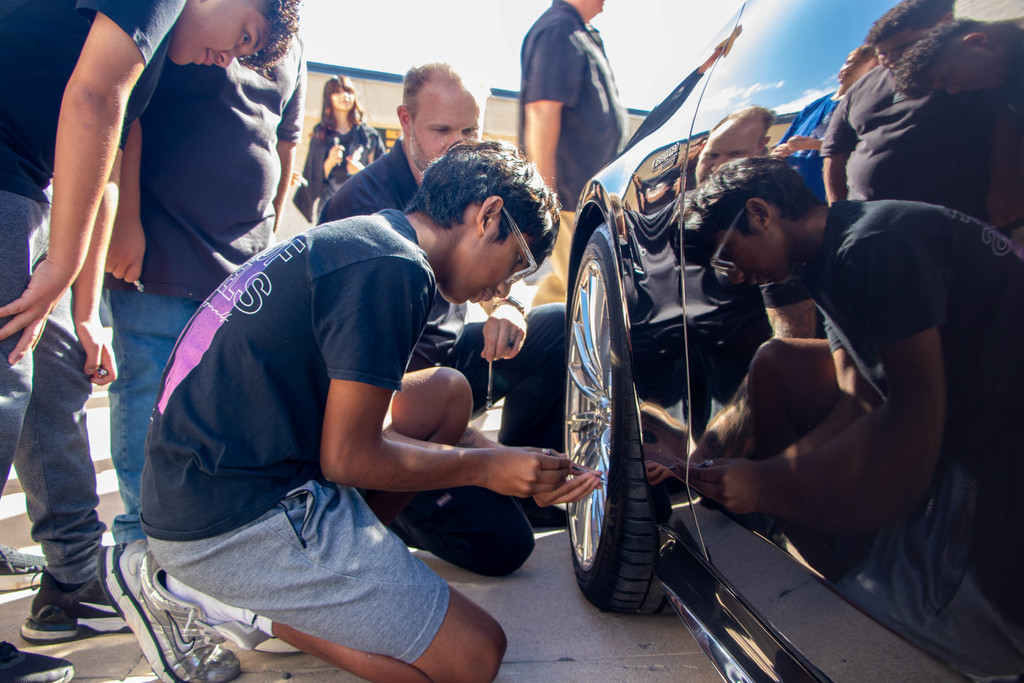 A student kneels beside a car using a tire gauge to check air pressure during an automotive skills activity. An instructor and classmates observe nearby.