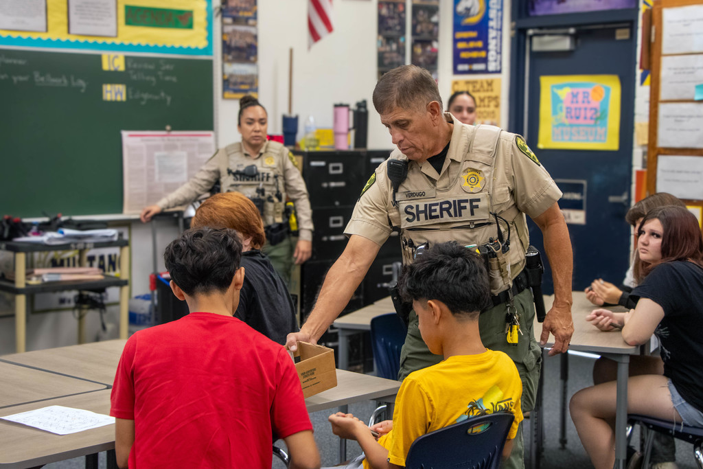 A Pima County Sheriff’s Department officer hands materials to students during a safety and crime prevention presentation in a classroom setting.
