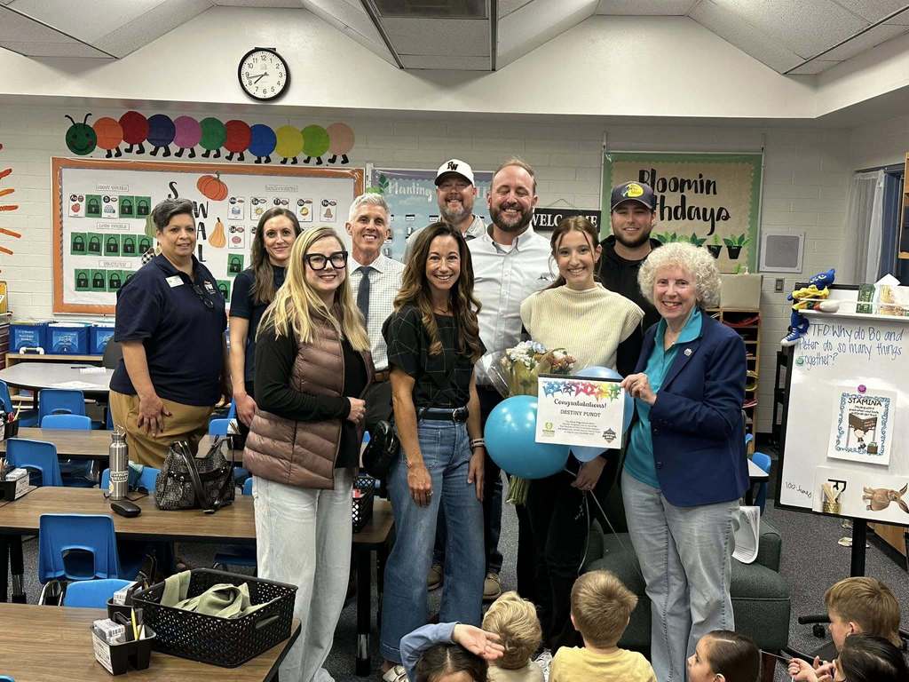 A group of adults stands in a kindergarten classroom celebrating Destiny Pundt’s Extra Yard for Teachers award. Destiny is holding flowers, balloons, and her certificate while smiling beside district and school staff. Young students are seated on the floor in the foreground watching the moment.