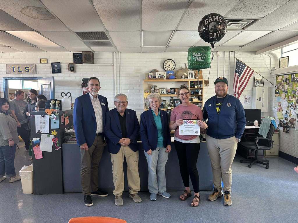 A group of adults stands inside a Flowing Wells High School classroom celebrating Mimi Lawton’s Extra Yard for Teachers award. Mimi is holding her certificate and smiling while staff members stand beside her. Balloons are displayed behind the group.