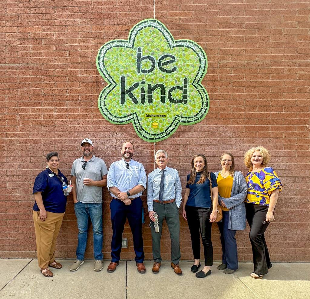 A group of seven adults stands smiling in front of a brick wall at Richardson Elementary. Behind them is a large green mosaic sign shaped like a flower that reads “be kind.”