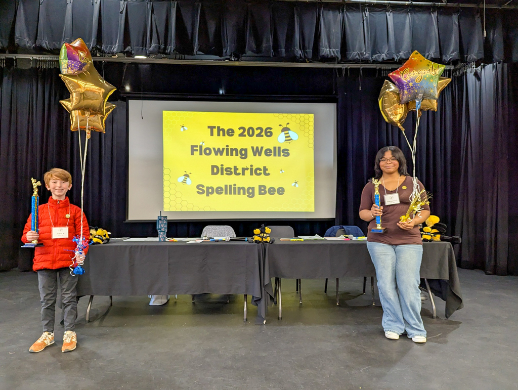 Two students stand on a stage holding trophies at the Flowing Wells District Spelling Bee. A large screen behind them reads “The 2026 Flowing Wells District Spelling Bee,” with balloons and decorations on each side.