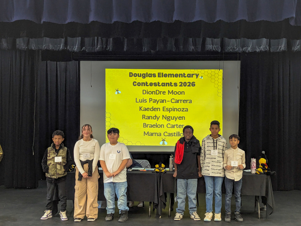 Douglas Elementary spelling bee contestants stand on stage with a screen behind them listing student names for the 2026 competition.