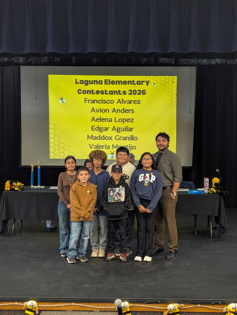 Laguna Elementary spelling bee contestants stand on stage with their teacher. A screen behind them displays the school name and student list for the 2026 competition.
