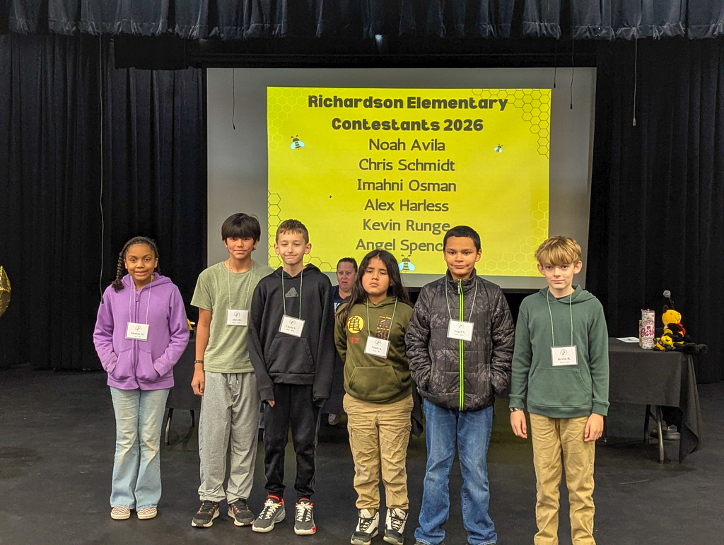 Richardson Elementary spelling bee contestants stand in a row on stage with a screen behind them showing the school name and student list for 2026.