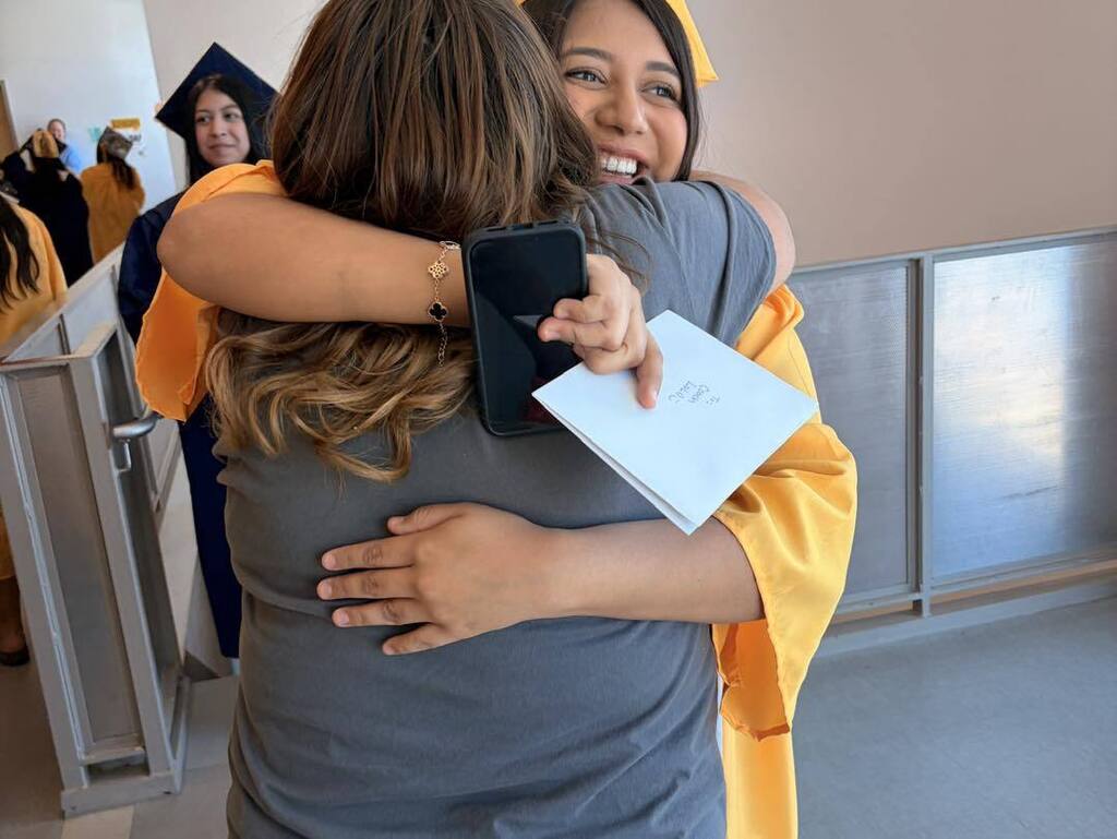 A graduating student in a gold cap and gown hugs a staff member or family member while holding a phone and card.