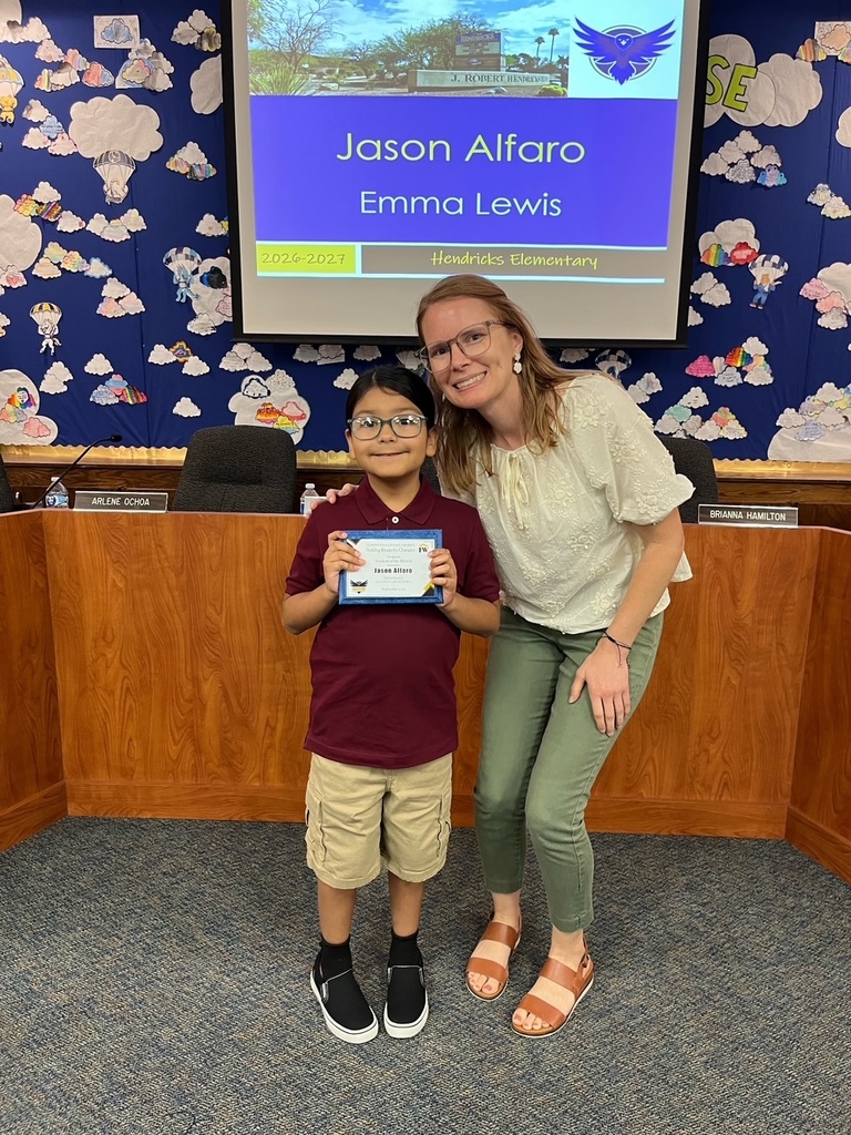 A student, Jason Alfaro stands beside an adult, Mrs. Lewis holding a “Building Blocks of Character” award certificate. They are smiling for a photo in front of a presentation screen at Flowing Wells School District.