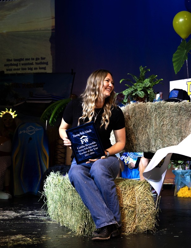 Flowing Wells High School teacher Caitlin Reynolds smiles while holding a plaque recognizing her as Teacher of the Year, seated on stage during opening assembly.