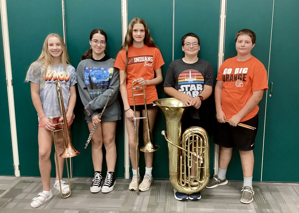 five middle school band students standing in front of a green wall with their instruments.