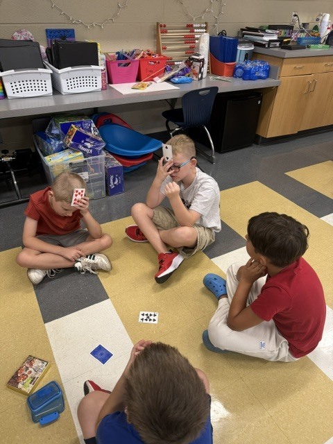 Four children sit on the floor, engaged in a card game with cards and books around them.