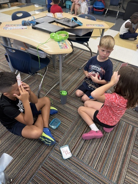 Three children in a classroom, seated on a carpeted floor, engaging in an activity.